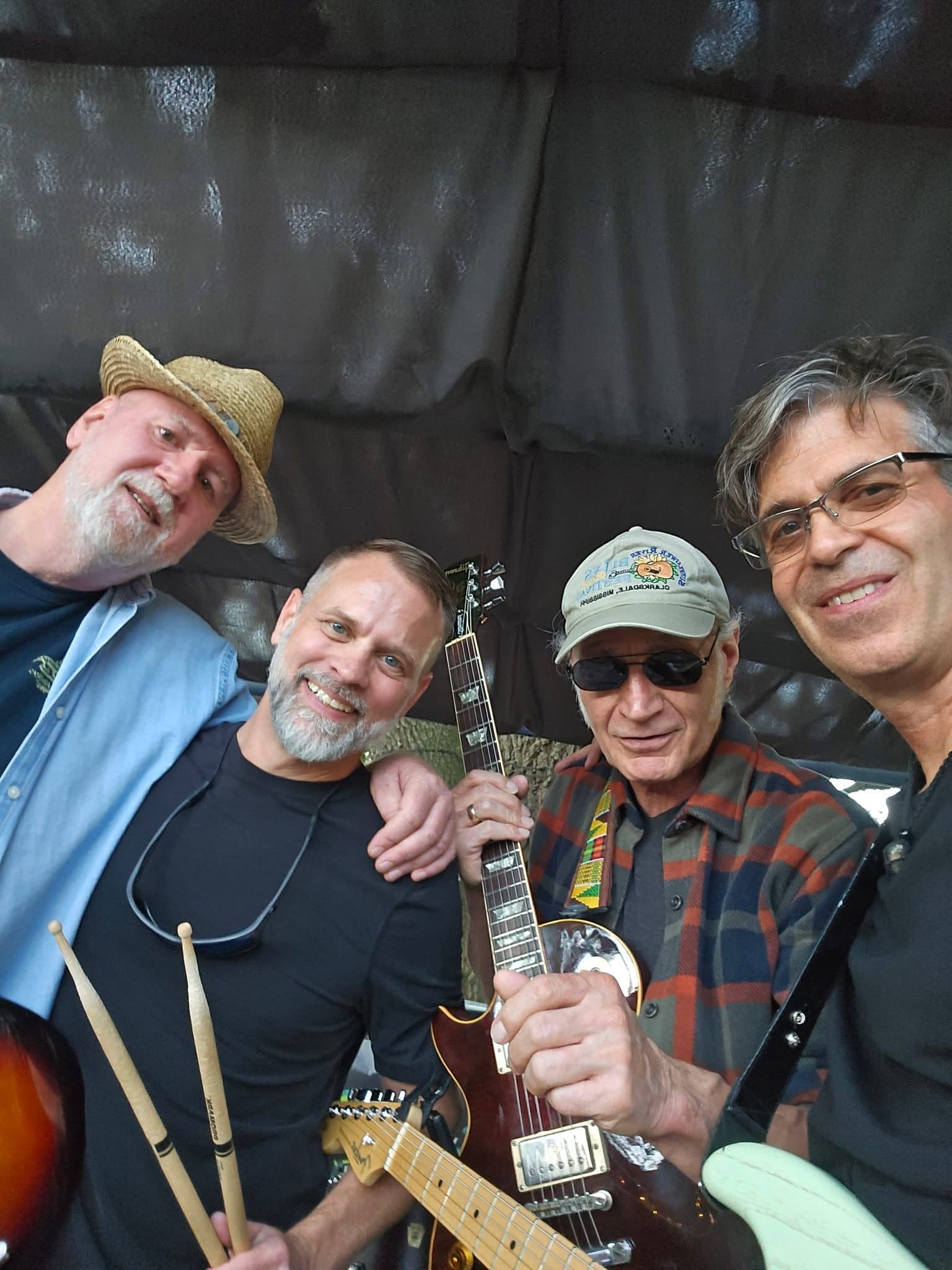 Close-up backstage selfie of the four band members with guitars and drumsticks