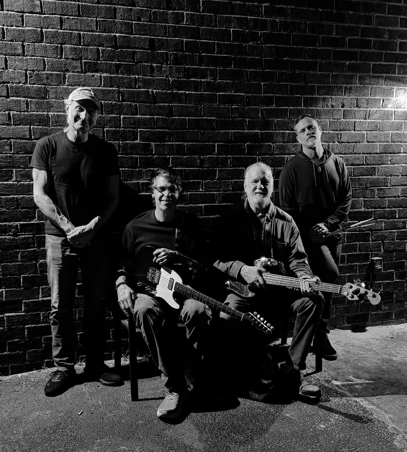 The four members of the Paul Stone Project with guitars against a brick wall, in black and white