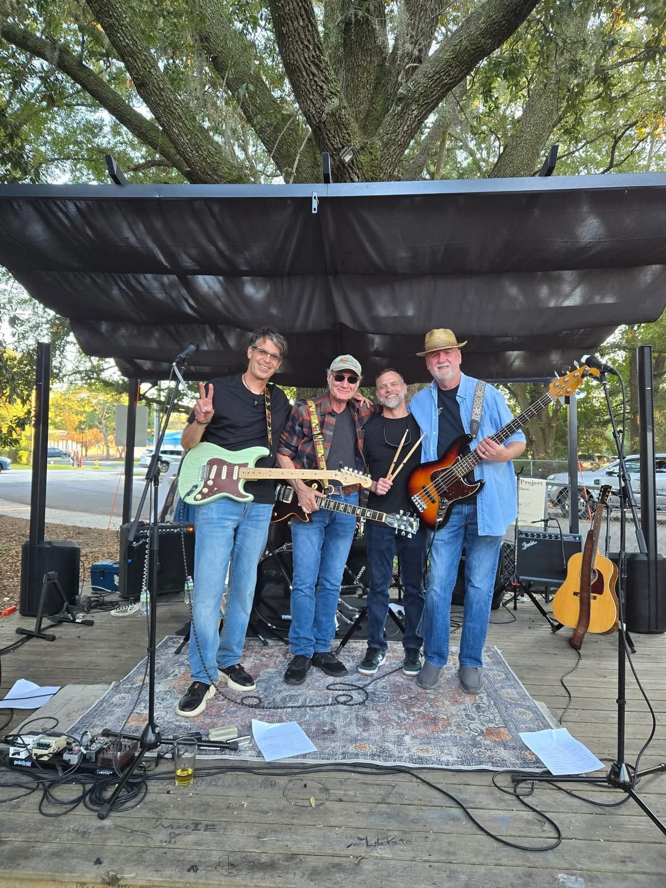The band posing together under an outdoor stage canopy beneath a large oak tree
